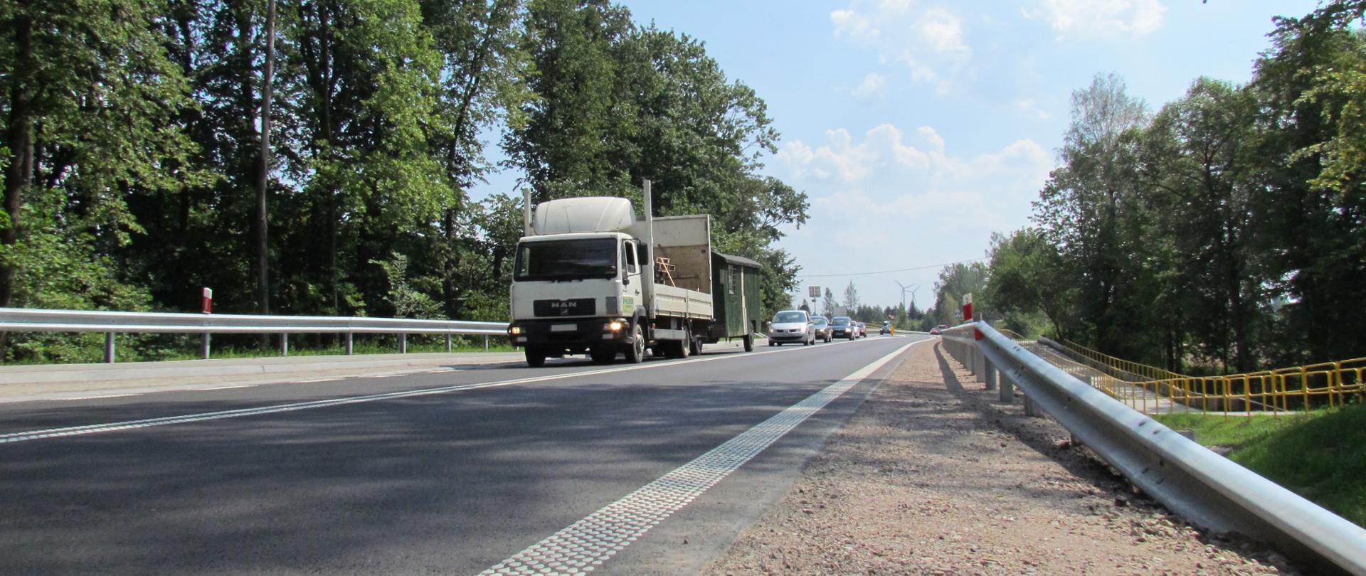 Asfaltowa droga, po obu stronach znajdują się bariery, za barierami drzewa. na drodze pojazdy osobowe i jeden ciężarowy.