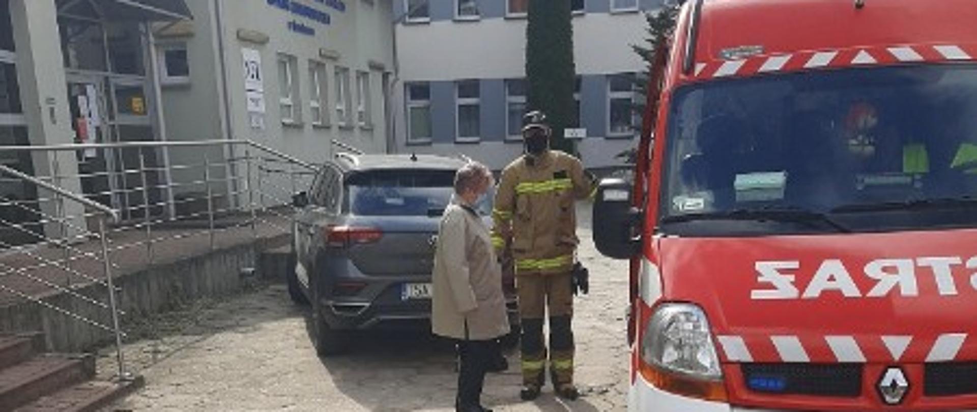 Zdjęcie zrobione przed budynkiem mieszkalnym. Strażak pomaga kobiecie wyjść ze strażackiego samochodu, którym była zawieziona na szczepienie.