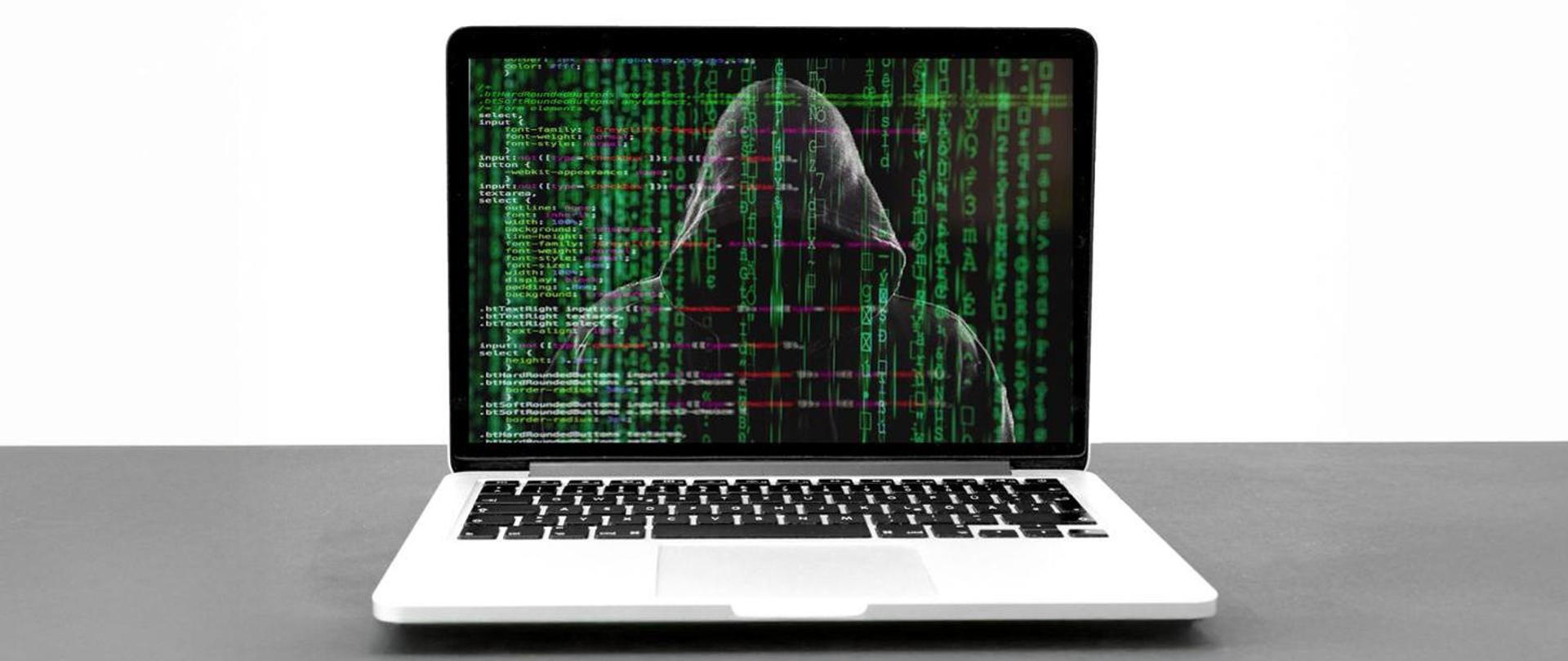 Zdjęcie przedstawia laptopa ustawionego na stole, na laptopie widać linijki kodu, w monitorze odbija się mroczna zakapturzona postać