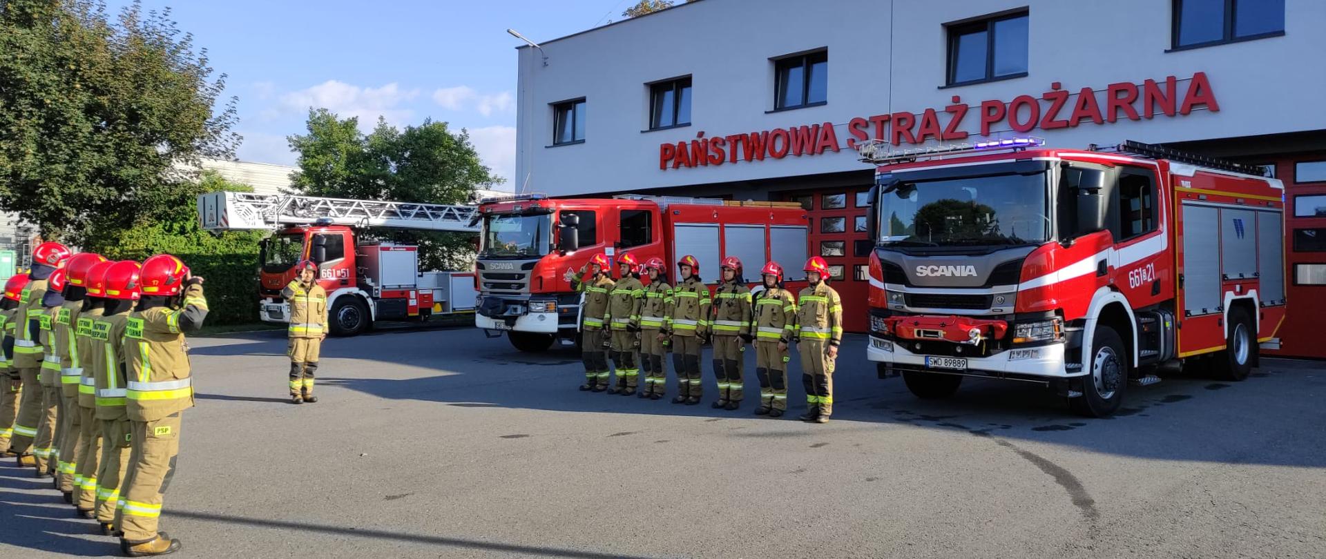 Na zdjęciu strażacy oraz samochody pożarnicze na tle komendy podczas uroczystej zbiórki