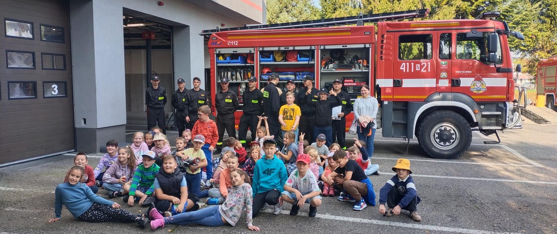 Dzieci na tle samochodu strażackiego, przy samochodzie strażacy.