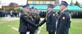 Rząd strażaków w umundurowaniu. Naprzeciwko dwóch funkcjonariuszy składają gratulacje i wręczają dokumenty strażakom stojącym w rzędzie.
