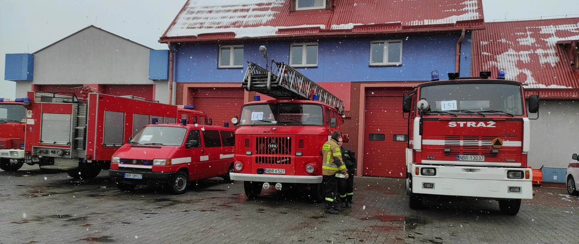 Cztery Czerwone samochody strażackie ustawione przy budynku. Pada śnieg.