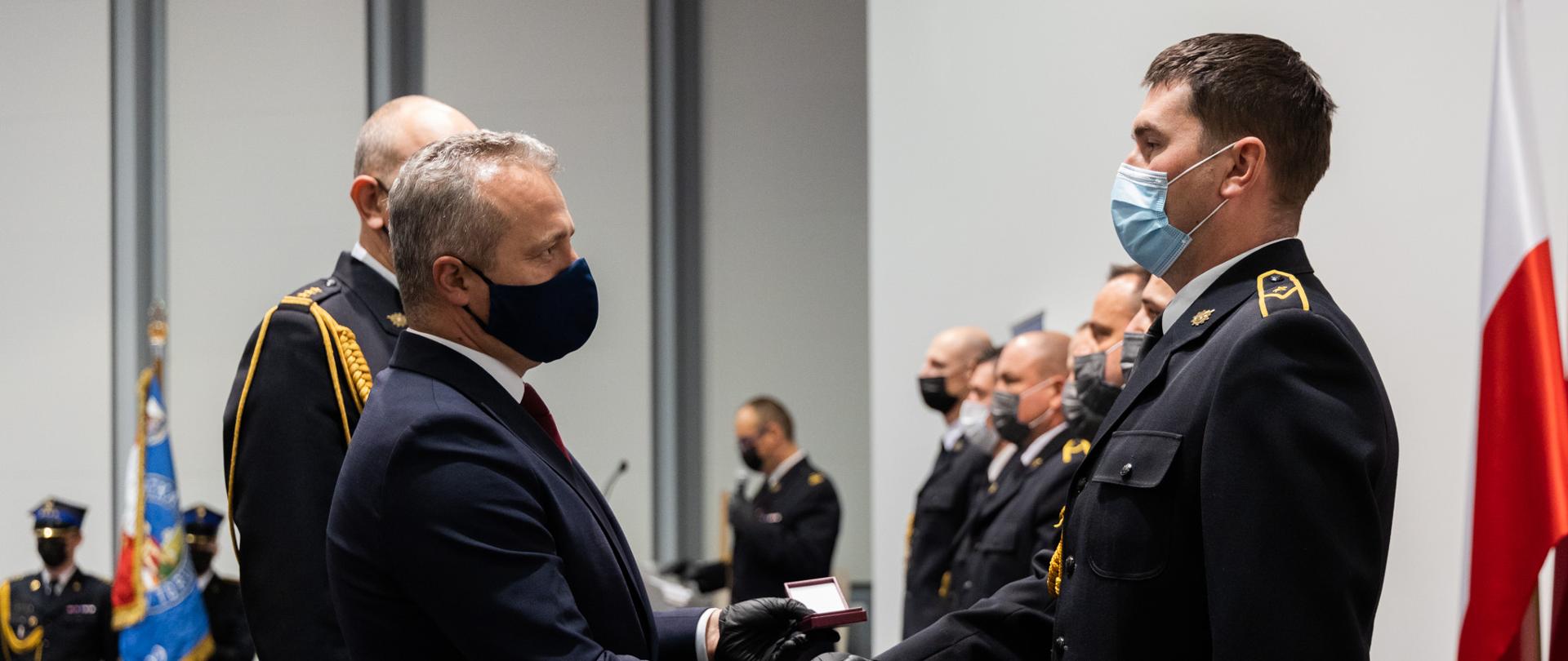 Na zdjęciu wyróżnieni strażacy ustawieni w szeregu. wojewoda jednemu z strażaków ściska dłoń i wręcza odznaczenia obok niego zastępca kujawsko-pomorskiego komendanta wojewódzkiego PSP, w tle stoi poczet sztandarowy.