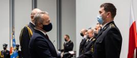 Na zdjęciu wyróżnieni strażacy ustawieni w szeregu. wojewoda jednemu z strażaków ściska dłoń i wręcza odznaczenia obok niego zastępca kujawsko-pomorskiego komendanta wojewódzkiego PSP, w tle stoi poczet sztandarowy.