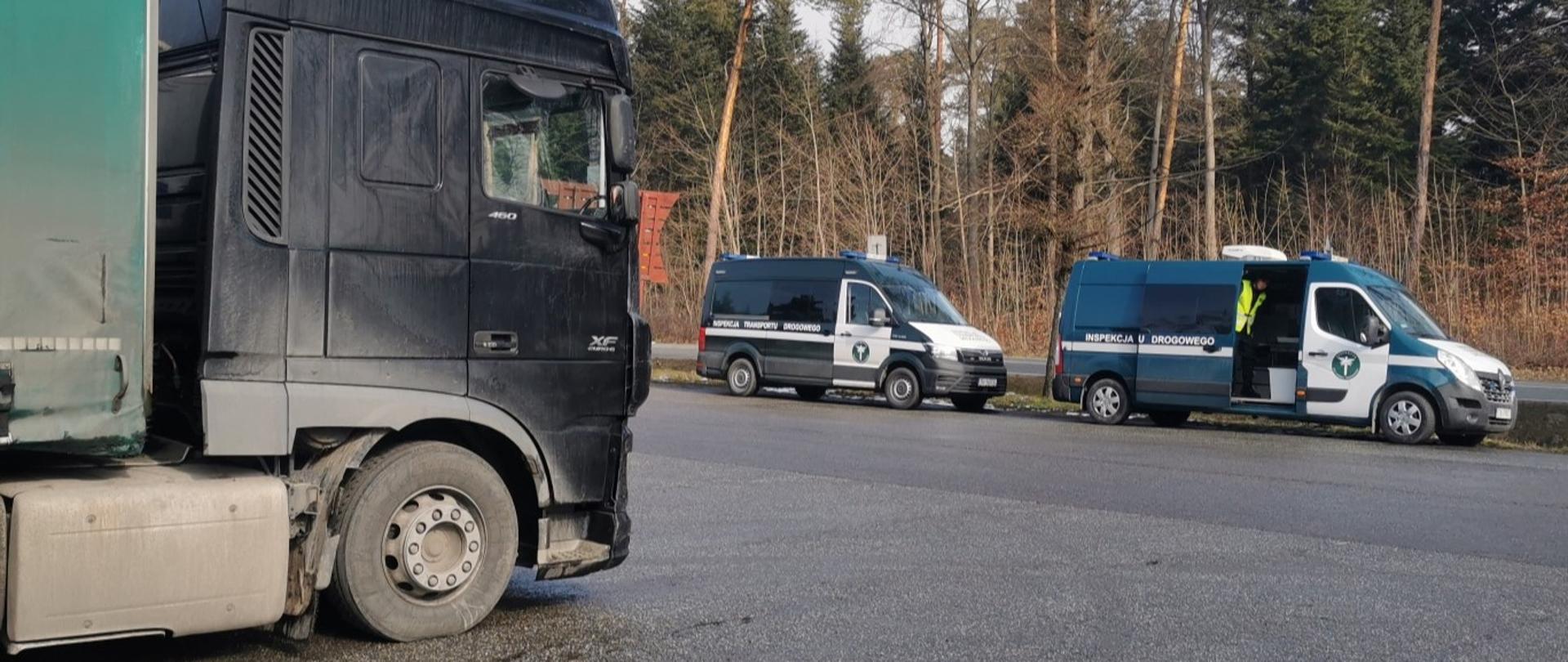 Na pierwszym planie (od lewej): bok ciągnika siodłowego z podpiętą naczepą typu firanka. W tle: w pobliżu zatrzymanego do kontroli zestawu ciężarowego stoją dwa oznakowane furgony świętokrzyskiej Inspekcji Transportu Drogowego, w tym jeden z odsuniętymi drzwiami bocznymi.