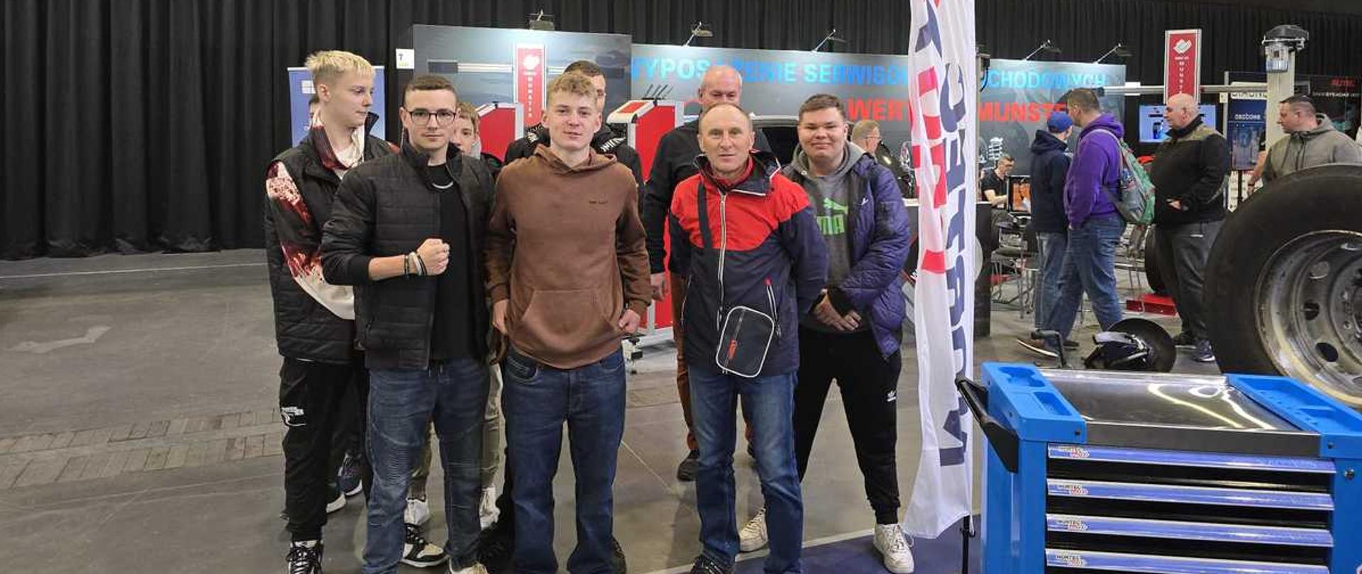 Zdjęcie przedstawia grupę siedmiu mężczyzn w różnym wieku, pozujących na tle hali wystawowej, prawdopodobnie podczas targów branżowych.
Oto szczegółowy opis:
Pierwszy plan: Grupa stoi na szarym, betonowym podłożu. Po prawej stronie widoczny jest fragment niebieskiego wózka narzędziowego z szufladami oraz duża opona ciężarowa. Obok nich stoi biała flaga reklamowa (tzw. wing) z czerwono-niebieskim logo.
Osoby:
Mężczyźni są ubrani w stroje codzienne: bluzy z kapturem, kurtki sportowe (w tym jedna jaskrawoczerwona) i jeansy.
Większość z nich patrzy bezpośrednio w stronę obiektywu, lekko się uśmiechając.
Tło:
Za grupą znajdują się stanowiska targowe z panelami informacyjnymi w kolorze niebieskim i czerwonym. Widoczne są napisy, m.in. fragmenty słów związanych z wyposażeniem serwisów.
W oddali, za stoiskami, widać czarną, pofałdowaną kurtynę, która oddziela przestrzeń wystawową.
Pod wysokim, przemysłowym sufitem zamontowane są liczne reflektory i lampy, które jasno oświetlają całą scenę.