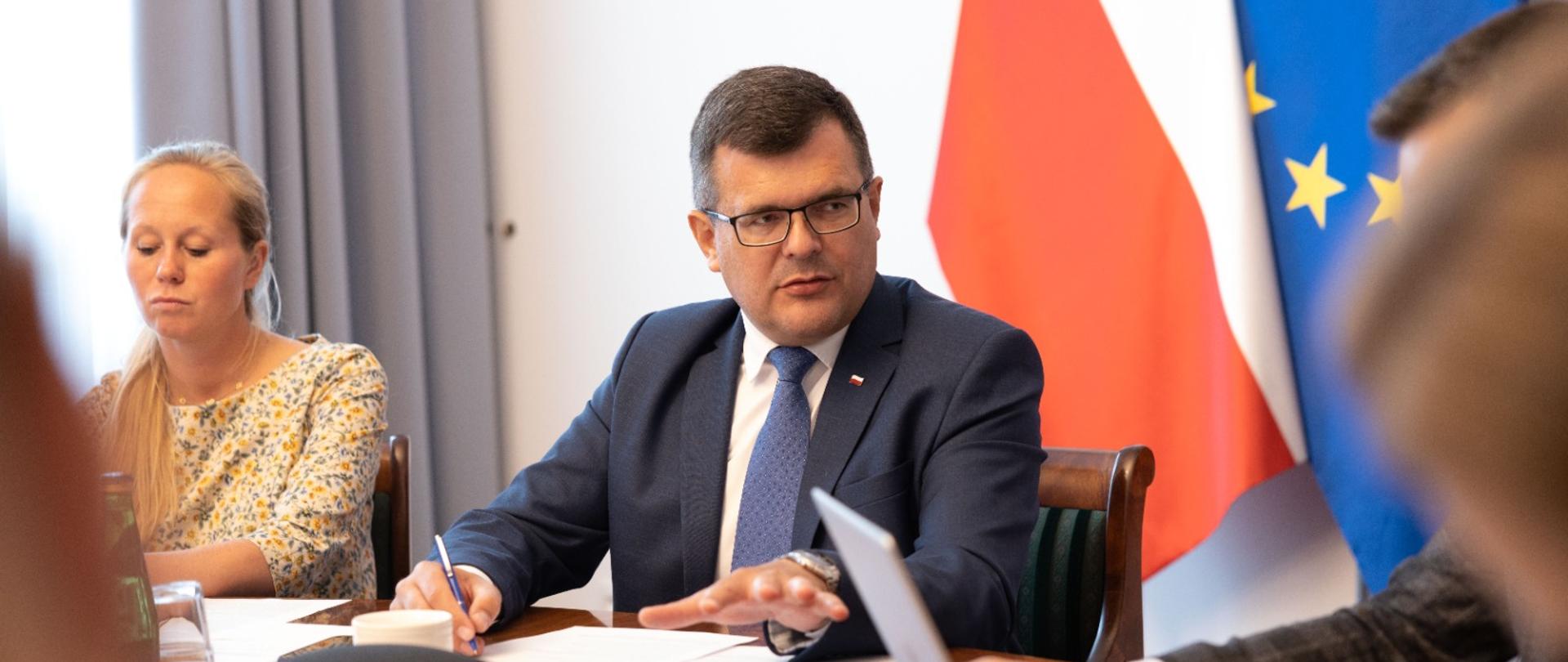 Przy okrągłym stole zasiadają uczestnicy posiedzenia, strona rządowa, pracodawców i pracobiorców. W oddali na środku fotografii, przewodniczący posiedzenia Pan Minister Piotr Uściński, a w tle za nim po dwie flagi: Polski i Unii Europejskiej oraz na ścianie godło Polski. Na stole znajdują się notatki oraz zastawa stołowa.