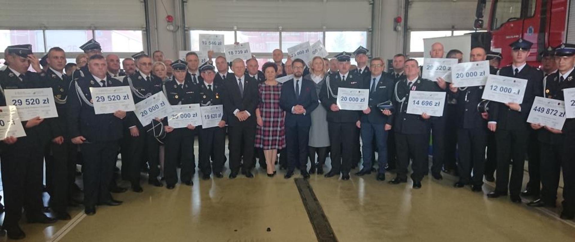 Zdjęcie wykonane w jednostce ratowniczo - gaśniczej Państwowej Straży Pożarnej w Radomiu. Na zdjęciu widać wydarzenie związane z podpisaniem umów na dofinansowanie jednostek ochotniczych straży pożarnych z terenu powiatu radomskiego, lipskiego, szydłowieckiego i zwoleńskiego ze środków wojewódzkiego funduszu ochrony środowiska i gospodarki wodnej. Wykonane zdjęcie jest zdjęciem grupowym. Na zdjęciu znajdują się dyrektor funduszu ochrony środowiska, parlamentarzyści, lokalni samorządowcy oraz przedstawiciele jednostek ochotniczych straży pożarnych. Przedstawiciele jednostek w rękach trzymają promesy na których znajdują się kwoty z przyznanymi dofinansowaniami. 