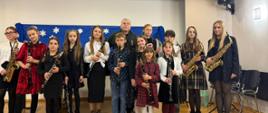 Grupa dzieci i młodzieży stoi na scenie w sali koncertowej, trzymając w rękach klarnety i saksofony. W środku grupy stoi nauczyciel. Wszyscy pozują do wspólnego zdjęcia po występie. Za nimi znajduje się niebieskie tło ozdobione białymi papierowymi śnieżynkami. Scena jest dobrze oświetlona, a uczestnicy są odświętnie ubrani.