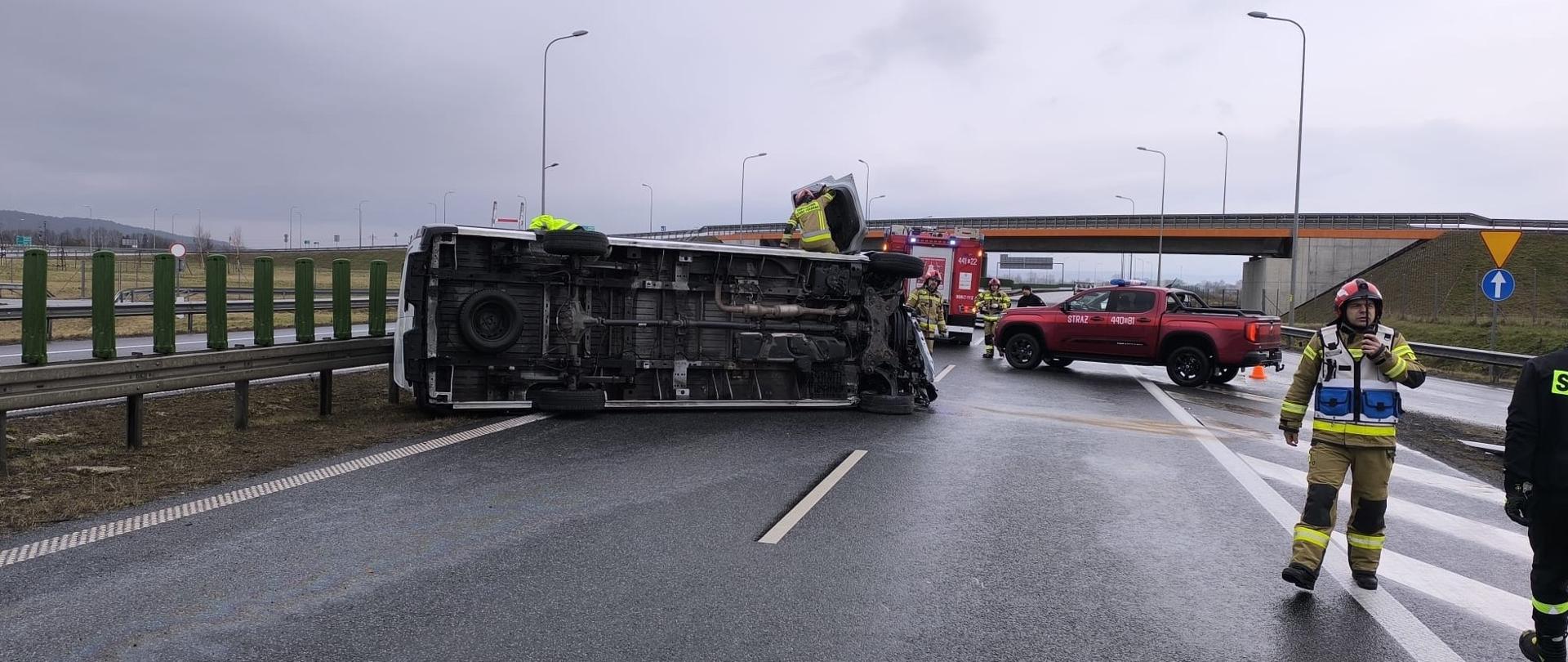 Na mokrej drodze szybkiego ruchu leży przewrócony na bok biały bus, częściowo blokując pas ruchu przy barierkach. Wokół pracują strażacy w odblaskowych ubraniach, a obok stoją czerwone pojazdy ratownicze zabezpieczające miejsce zdarzenia. Na niebie wiszą ciężkie chmury, a cała scena przedstawia działania służb podczas usuwania skutków wypadku drogowego.