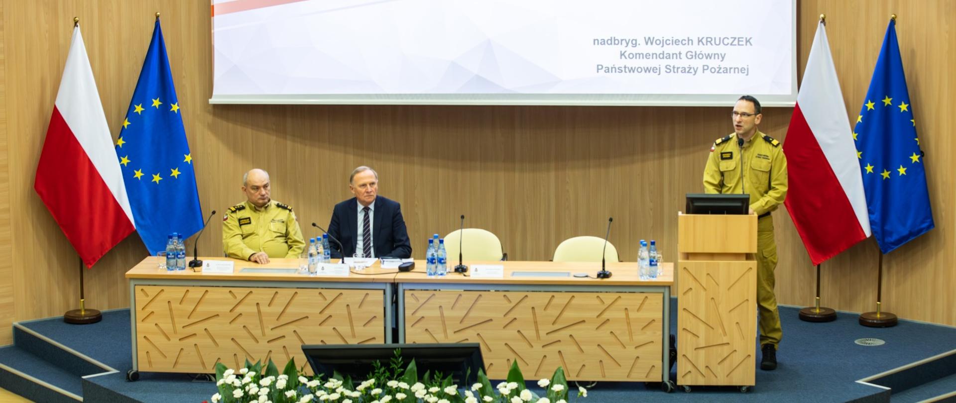 Sala konferencyjna Państwowej Straży Pożarnej. Przy stole prezydialnym siedzą przedstawiciele kierownictwa PSP oraz zaproszony gość, a jeden z oficerów PSP przemawia z mównicy. W tle widoczny ekran z prezentacją oraz flagi Polski i Unii Europejskiej.