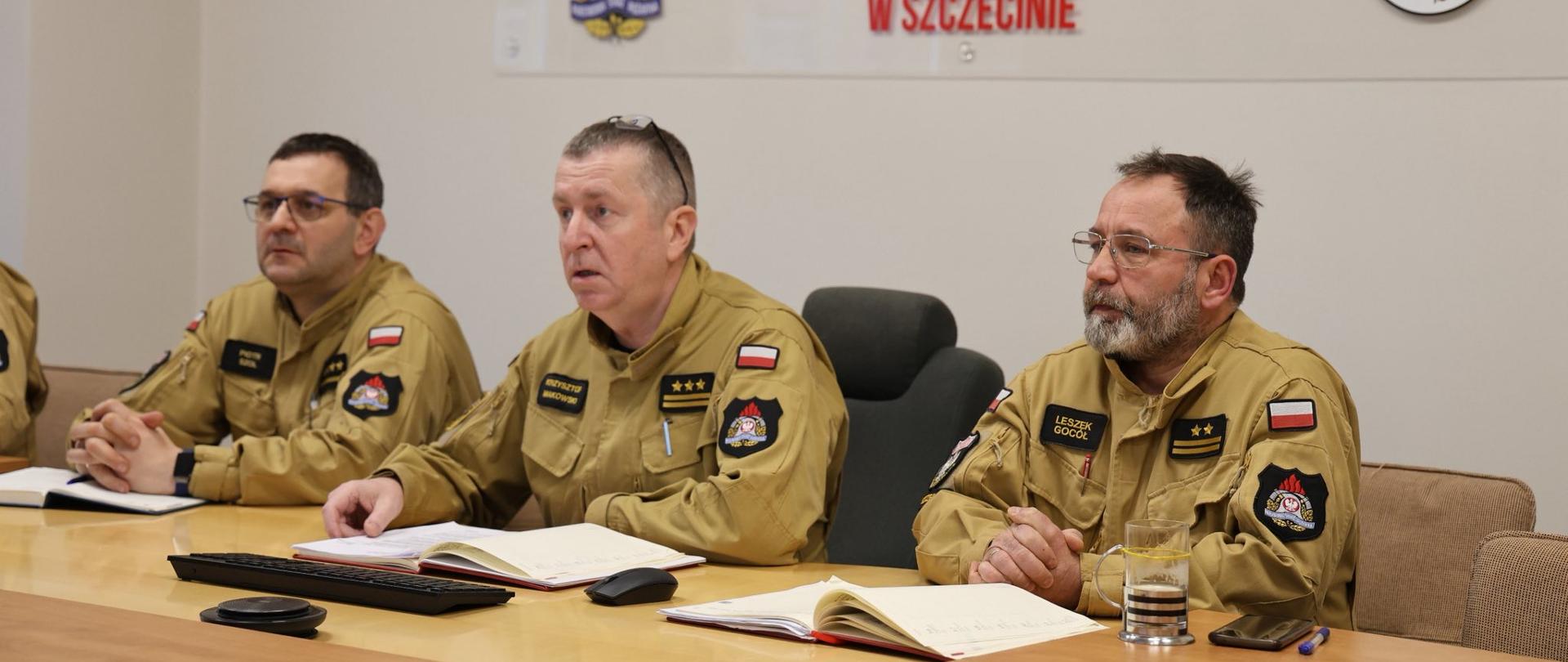 Na zdjęciu widać trzech mężczyzn w mundurach Państwowej Straży Pożarnej siedzących przy stole w sali konferencyjnej. Przed nimi leżą otwarte notatniki i dokumenty. W tle znajduje się tablica z napisem „Komenda Wojewódzka Państwowej Straży Pożarnej w Szczecinie” oraz godła.