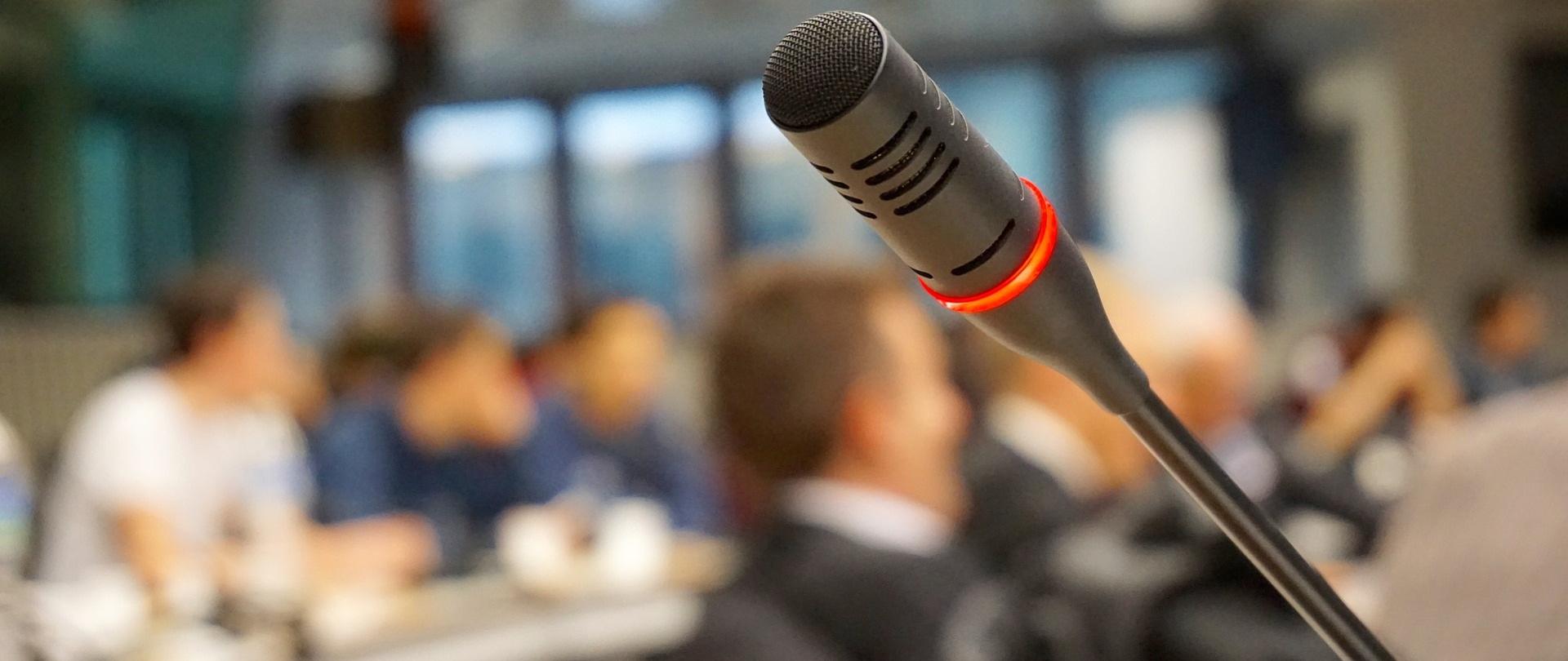 mikrofon, w tle grupa ludzi siedzących za stołami
