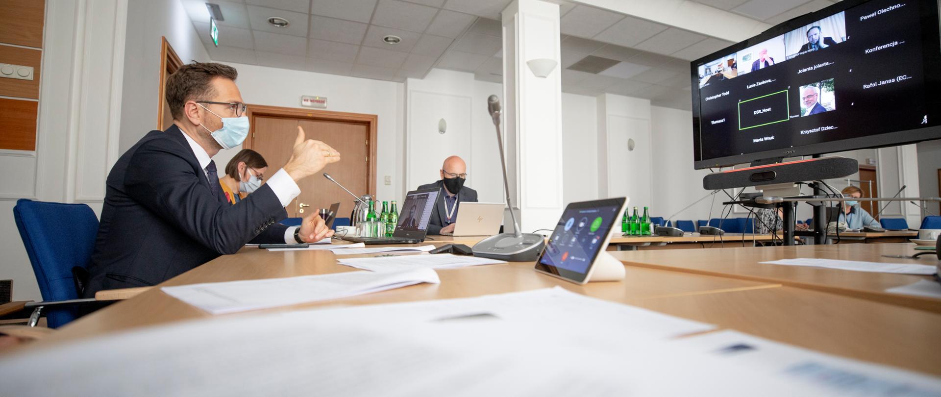 Minister Buda siedzi przy stole przed nim stoi telewizor, na którym trwa wideokonferencja 