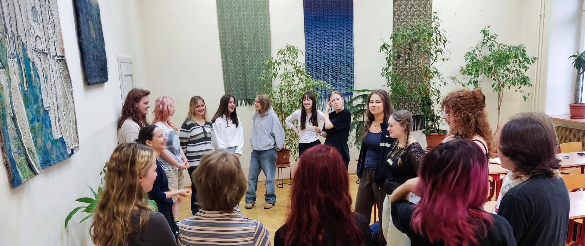 Grupa młodych osób stoi w kręgu w jasnej sali z dekoracjami na ścianach i roślinami w tle.