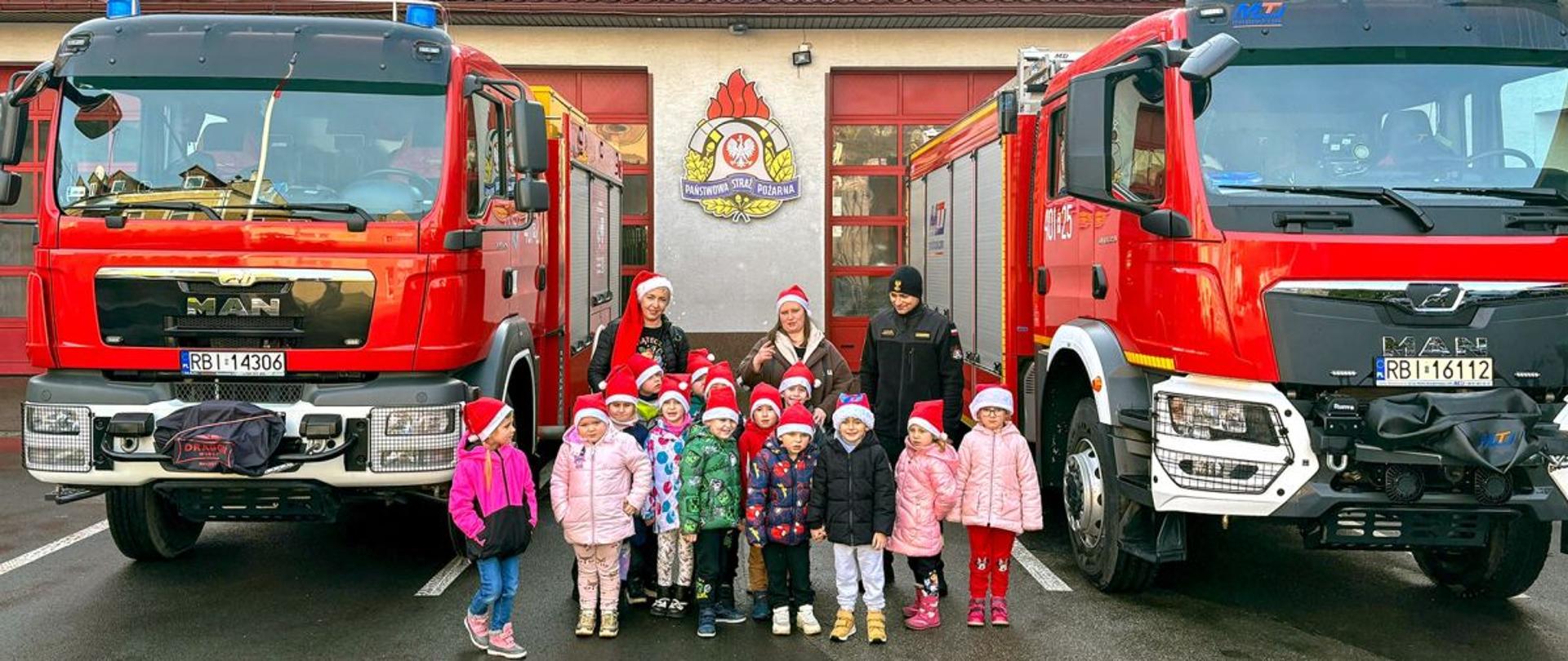 Na zdjęciu widać dzieci, dwie nauczycielki i strażaka przy samochodach pożarniczych. Dzieci i nauczycielki mają na głowach czapki św. Mikołaja. W tle garaże JRG Ustrzyki Dolne. 