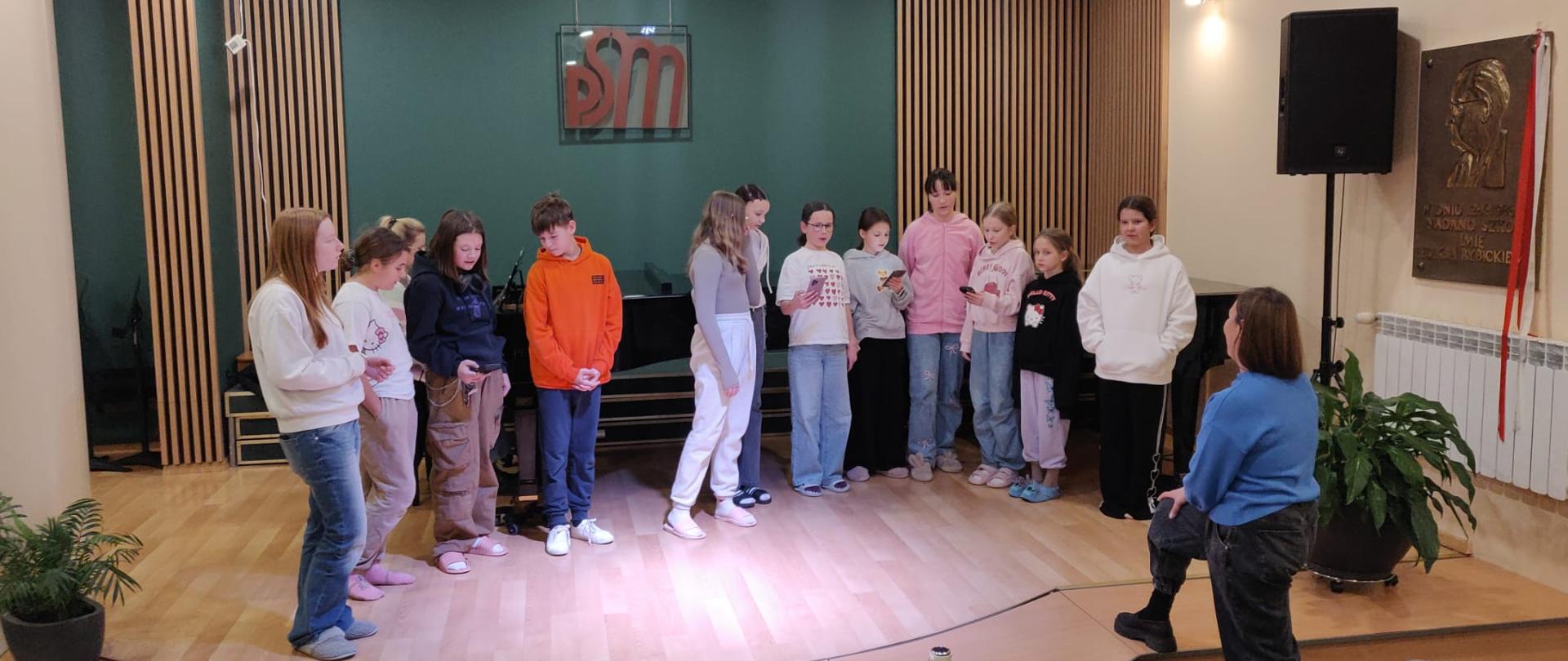 Dzieci prezentują się na scenie w trakcie warsztatów wokalnych są w luźnych codziennych ubraniach w tle widoczna jest Zielona ściana oraz Czarny fortepian.