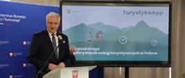 Wicepremier, minister rozwoju, pracy i technologii Jarosław Gowin stojący przy mównicy z mikrofonem, po jego prawej stronie ekran telewizora z wyświetloną grafiką przedstawiającą dwóch turystów w górach z plecakami i napisem TurystykApp - najlepsza droga do odkrywania usług turystycznych w Polsce.