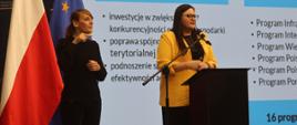 Minister funduszy i polityki regionalnej Małgorzata Jarosińska-Jedynak stoi przy pulpicie i przemawia do mikrofonu, za jej plecami grafika z prezentacją, po lewej stronie stoi tłumaczka języka migowego