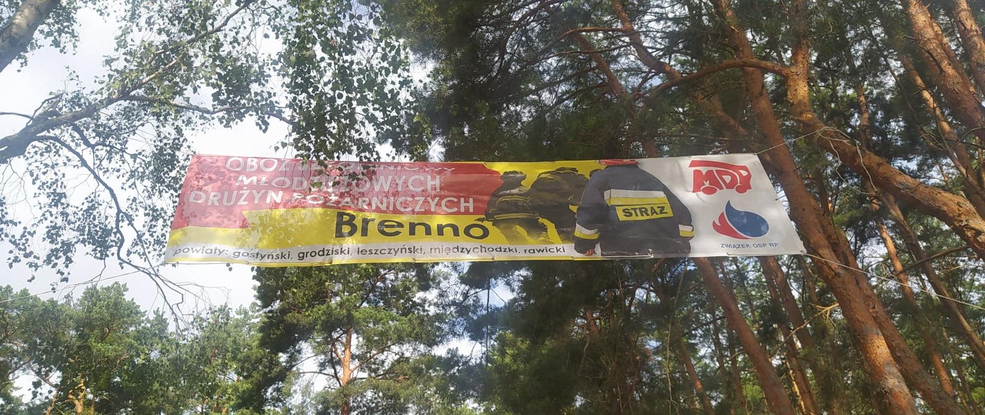 Obóz szkoleniowo-wypoczynkowy MDP w Brennie. Pomiędzy drzewami zawieszony jest kolorowy baner, na którym znajduje się napis: " OBÓZ SZKOLENIOWY MŁODZIEZOWYCH DRUŻYN POŻARNICZYCH BRENNO". POd napisem wymienione są powiaty, z których młodzież uczestniczy w obozie. Z prawej strony sylwetki strażaków, a obok nich logo MDP i ZOSP RP.