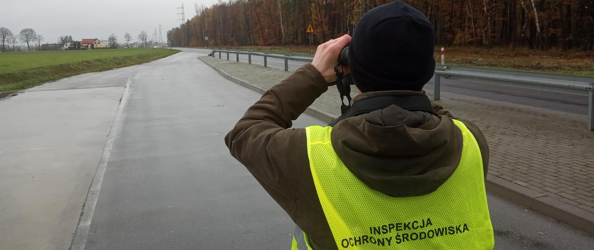 Inspektor ubrany w czapkę, ciemną kurtkę oraz będący w kamizelce z napisem Inspekcja Ochrony Środowiska stoi na poboczu drogi obserwując przez lornetkę sytuację w oddali. Po lewej stronie pole, po prawej stronie las.