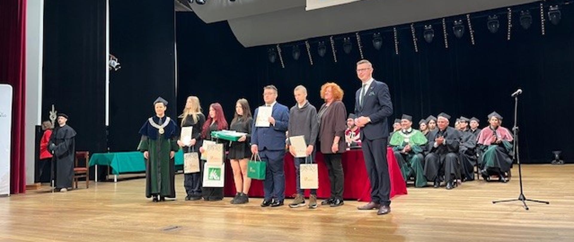 na pierwszym planie stoi osiem osób w głębi sceny siedzi duża grupa