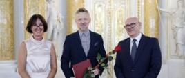 Na tle bogato zdobionych drzwi otoczonych kolumnami i posągami stoją trzy uśmiechnięte osoby. W środku nowo wybrany dyrektor z listem nominacyjnym i czerwoną różą w dłoni. Po prawej stronie mężczyzna będący urzędnikiem ministerstwa. Po lewej stronie kobieta – urzędniczka ministerstwa.