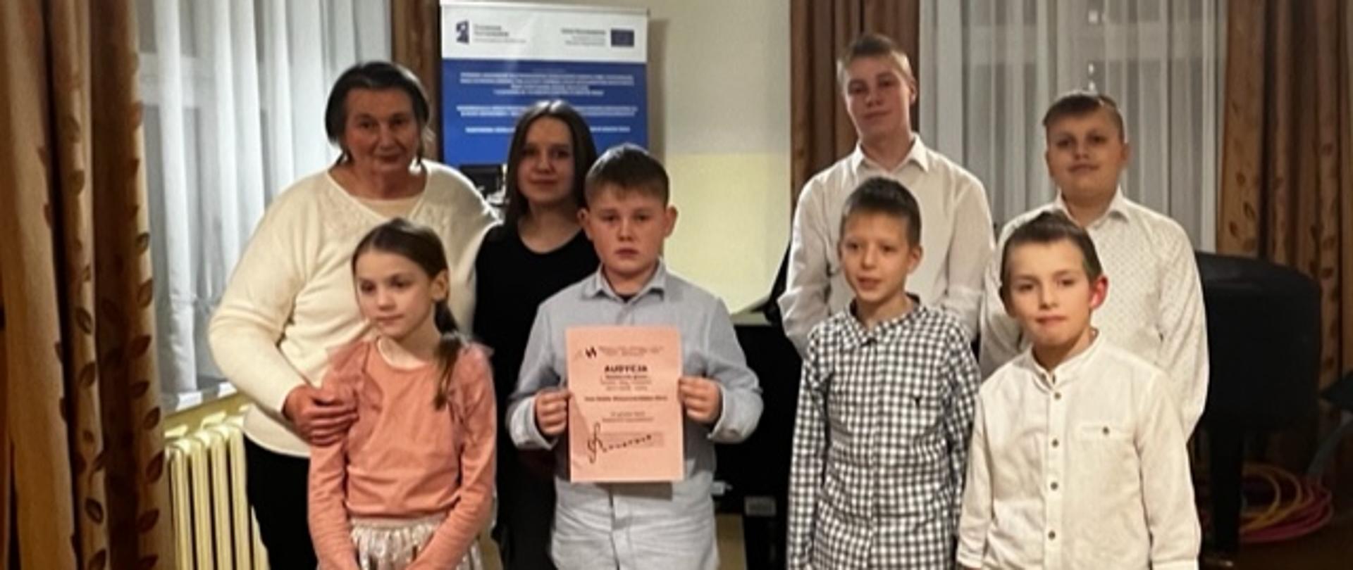 Zdjęcie grupowe uczniów biorących udział w audycji oraz nauczycielki instrumentu głównego. Uczestnicy stoją w dwóch rzędach jeden z uczniów trzyma plakat informujący o wydarzeniu. Za nimi dwa okna przysłonięte firanami i zasłonami. Pod oknem kaloryfer. Za nimi również fortepian oraz baner unijny. Od sufity zwisają dwie lampy zapalone.