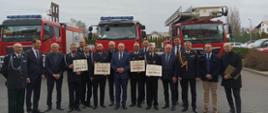 Zdjęcie przedstawia komendanci powiatowego PSP, przedstawiciele władz samorządowych oraz przedstawicieli jednostek OSP, które otrzymały promesy na tle trzech strażackich samochodów.