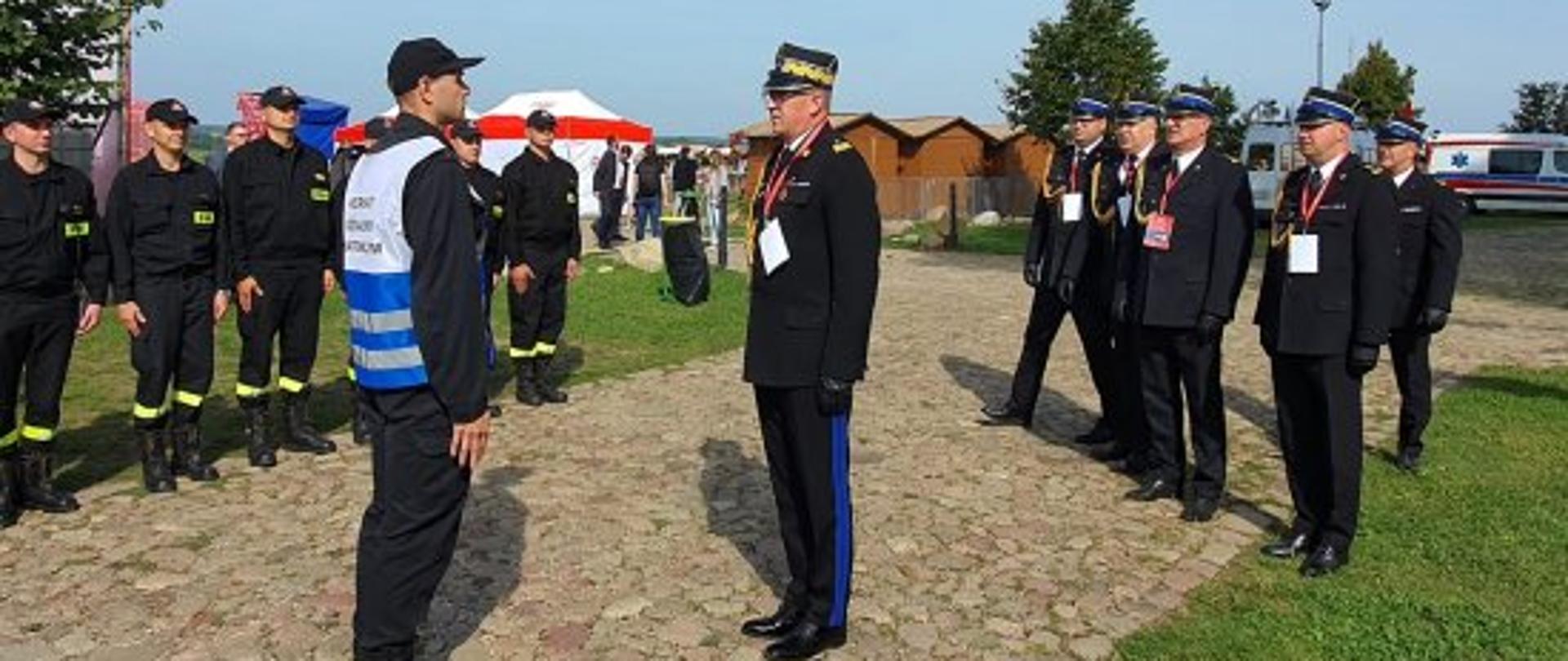 grupa strażaków stojących na bruku, dwóch strażaków stoi na przeciwko siebie, w tle karetka pogotowia oraz namioty