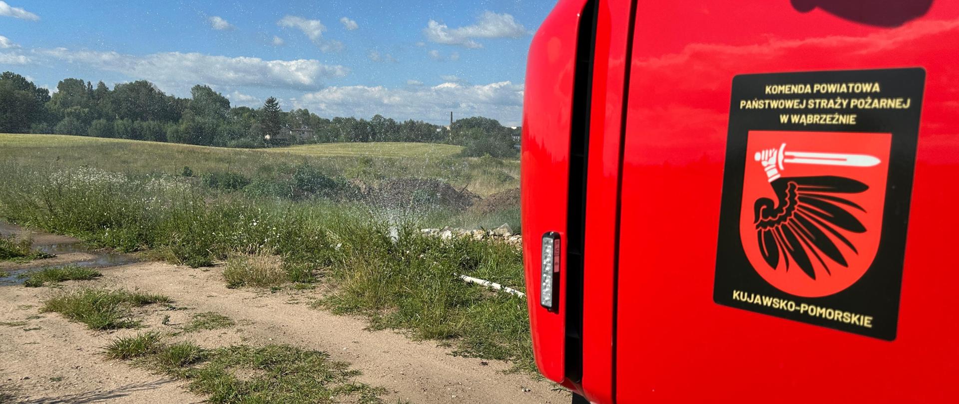 Fragment drzwi pojazdu pożarniczego z logo Komendy Powiatowej PSP w Wąbrzeźnie. Przed pojazdem znajduje się zakończenie linii wężowej kurtyną wodną, z której wydobywa się woda.