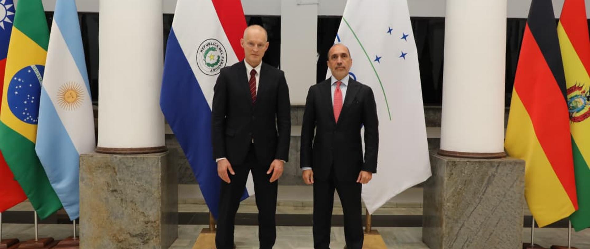 Na zdjęciu jest dwóch mężczyzn w garniturach. Patrzą w obiektyw. Za nimi są flagi.