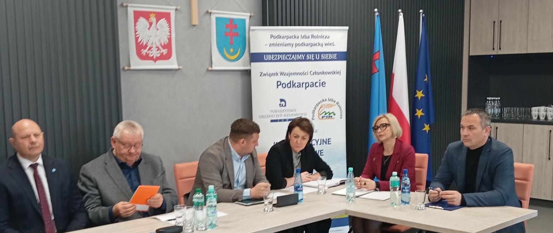Uczestnicy spotkania podczas dyskusji przy stole konferencyjnym; część osób analizuje dokumenty i materiały, a w tle widoczne są flagi oraz oznaczenia instytucji współorganizujących wydarzenie.