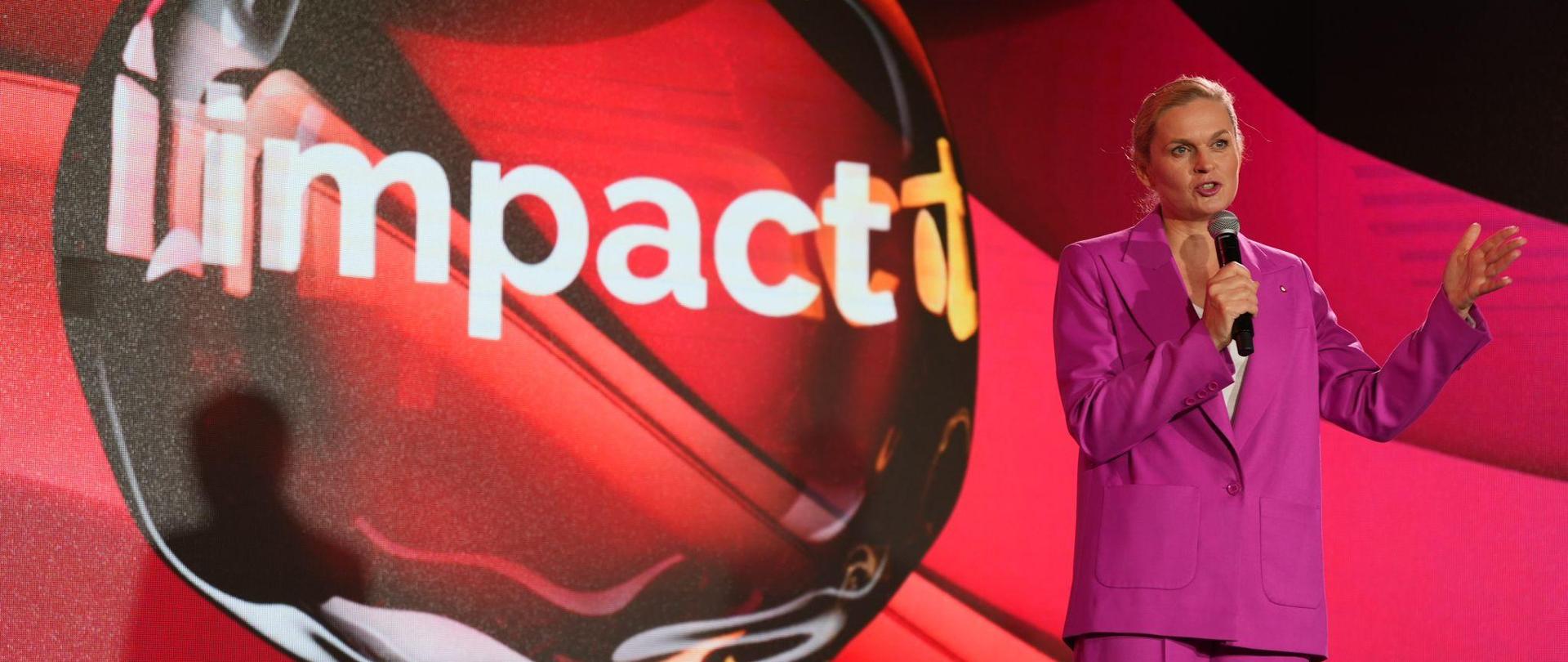 minister na stojąco przemawia do mikrofonu do uczestników wydarzenia. Ubrana w różowy garnitur. W tle logo na czerwonym tle z hasłem Impact. Impact 2024 z udziałem minister Barbary Nowackiej 