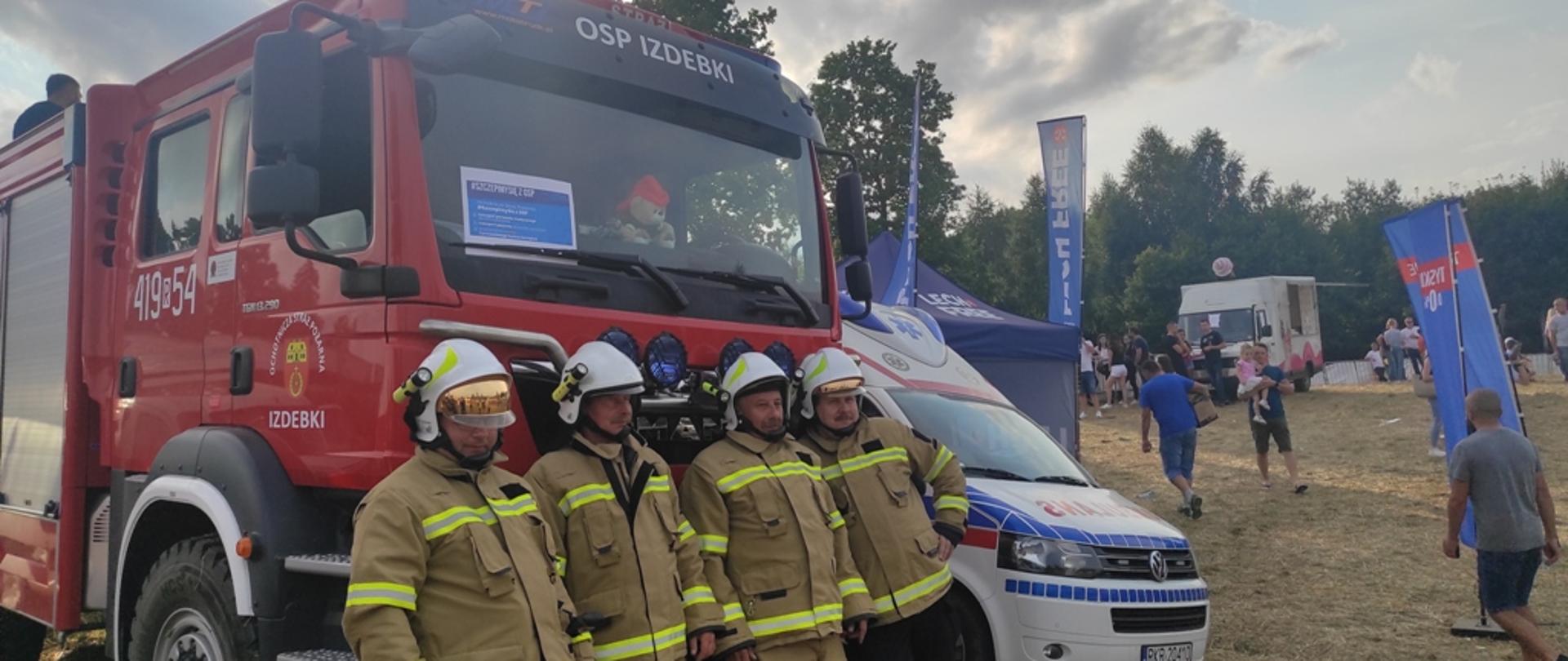 Kolorowe zdjęcie wykonane w przestrzeni otwartej w dzień. Przedstawia na środkowym planie przód czerwonego pojazdu pożarniczego i stojących przed nim strażaków. Są umundurowani na bojowo w kolorze piaskowym i w białych hełmach na głowach. Na samochodzie widoczne białe napisy: OSP Izdebki a za szybą pojazdu kartka z napisem Szczepimy się z OSP na niebieskim tle. Z tyłu widoczna częściowo karetka medyczna, namioty stoiskowe, inne pojazdy i cywilne osoby. W tle widoczne zielone drzewa.