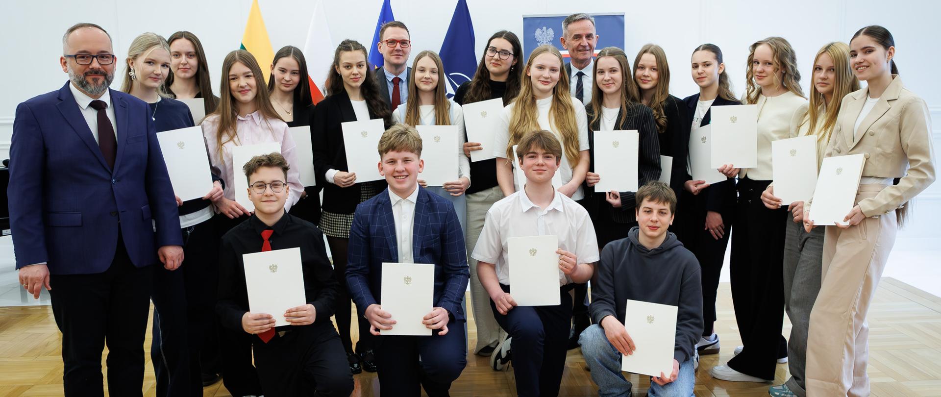 Zdjęcie przedstawia grupę około dwudziestu kilku osób ustawionych w dwóch rzędach w jasnym, przestronnym pomieszczeniu o drewnianej podłodze. Wszyscy stoją lub kucają blisko siebie, tworząc zwartą grupę. Każda osoba trzyma w dłoniach jasny, oficjalny dokument. W tle, na środku i po bokach, ustawione są flagi na stojakach. Osoby w grupie mają na sobie eleganckie lub półformalne stroje.