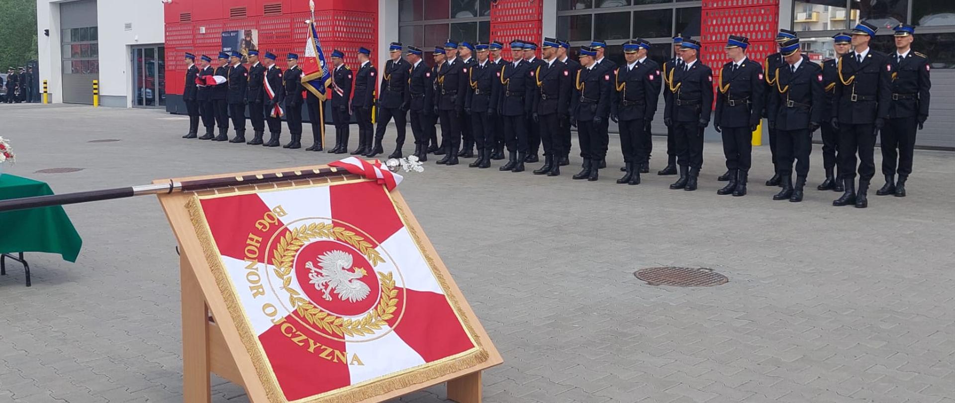 Zdjęcie sztandaru na tle jednostki ratowniczo-gaśniczej. Przed jednostką stoją w mundurach strażacy.