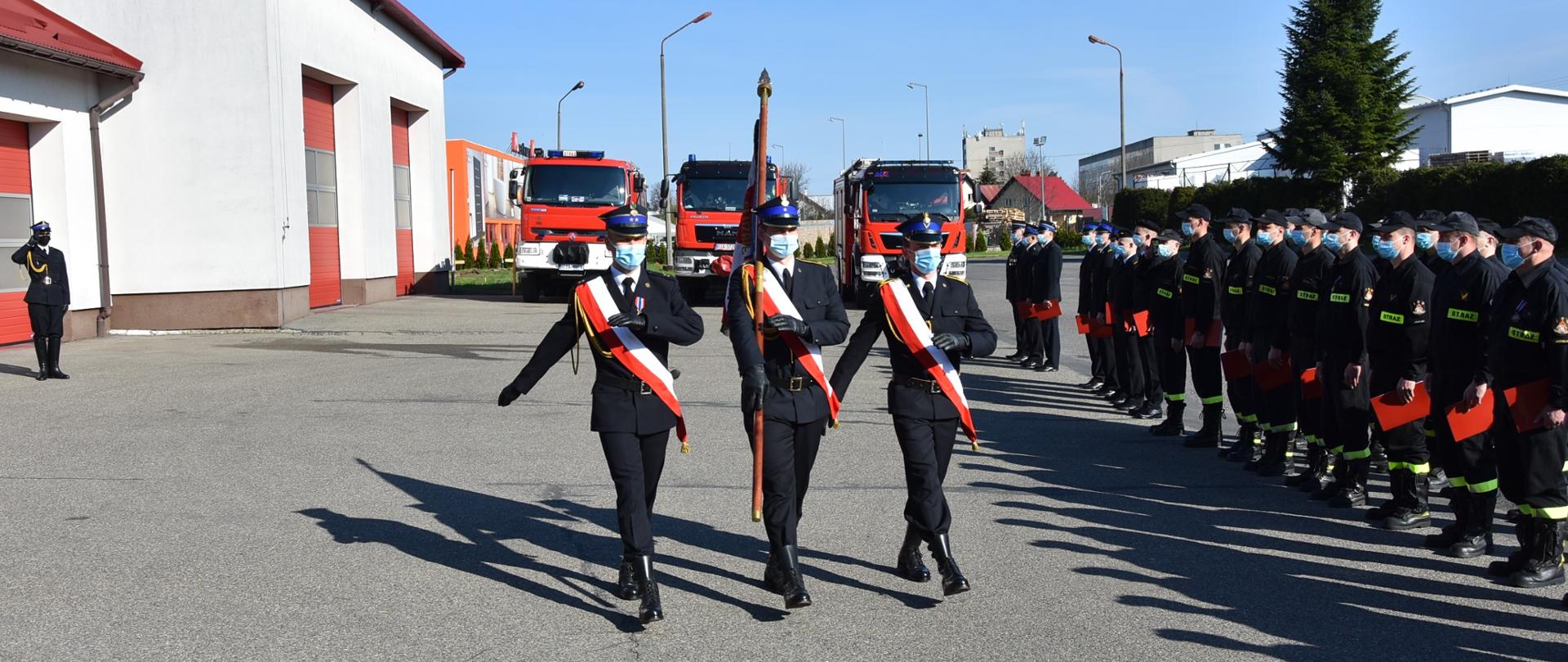 Na zdjęciu na pierwszym planie widać maszerujący poczet sztandarowy. Po prawej stronie widać strażaków stojących w dwuszeregu, a z tyłu trzy wozy strażackie. Wszystkie osoby posiadają maseczki ochronne.
