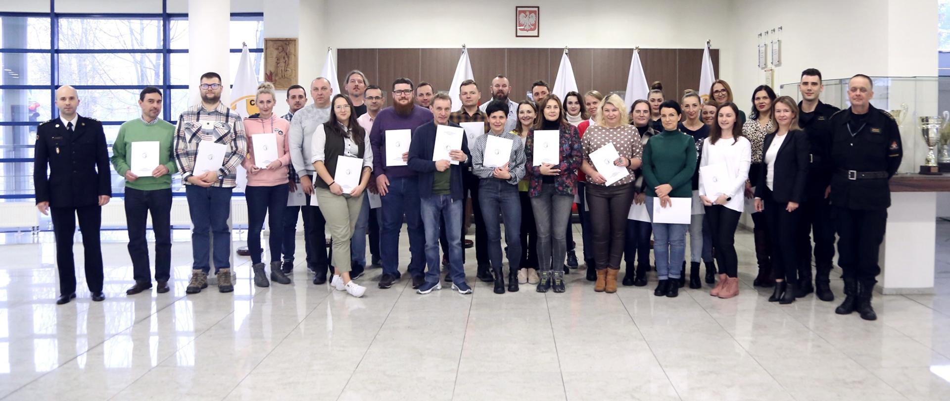 Grupowe zdjęcie uczestników szkolenia (28 osób) w towarzystwie Zastępcy Komendanta Centralnej Szkoły PSP, dwóch funkcjonariuszy CS PSP oraz kobiety. Uczestnicy szkolenia trzymają otrzymane zaświadczenia