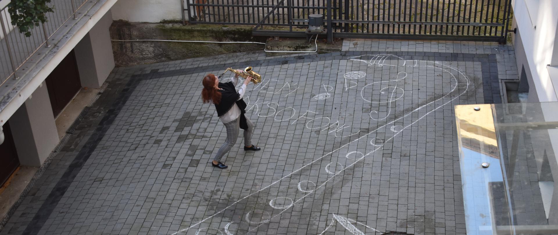 Zdjęcie przedstawia nauczyciela z saksofonem w dłoniach na dziedzińcu szkoły, na którym narysowany jest saksofon oraz napis Dzień Saksofonu. Kolorystyka zdjęcia jest szara. 