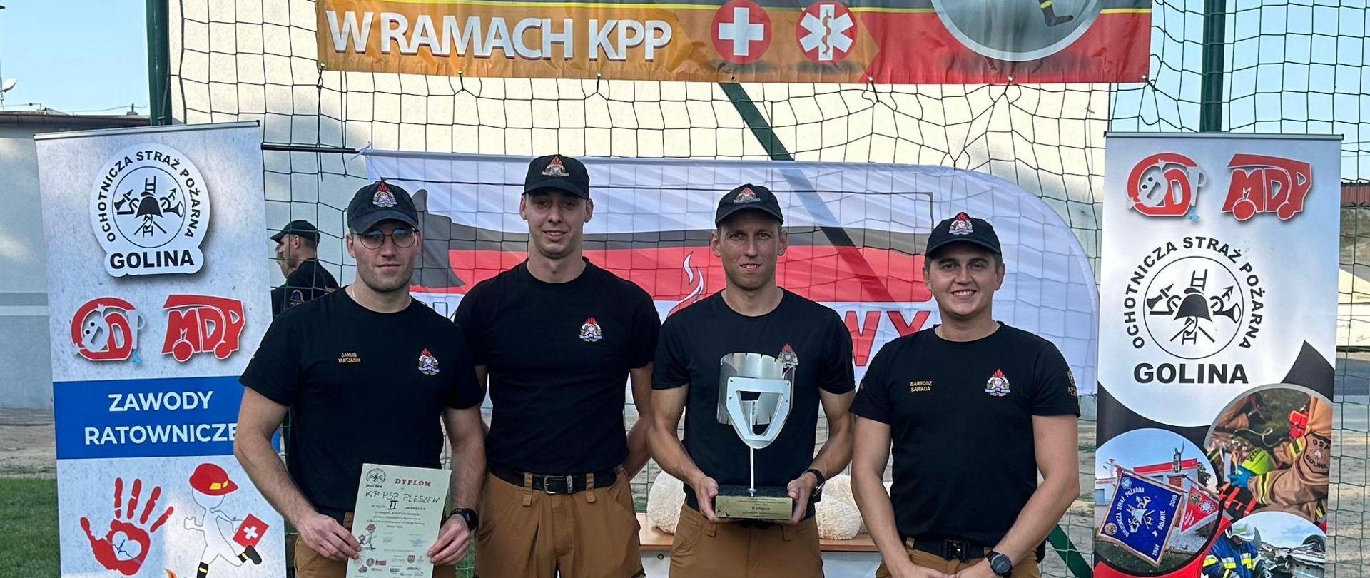 Czterech mężczyzn w mundurach Państwowej Straży Pożarnej stoi w szeregujeden z nich trzyma duży srebrny puchar, kolejny trzyma w ręku dyplom, następny trzyma dużą papierową torbę