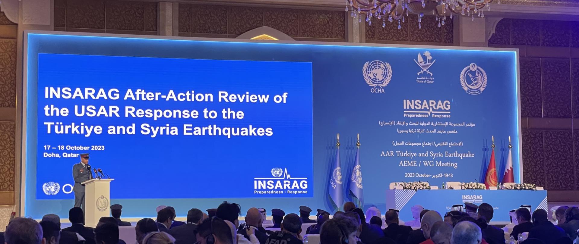 Na zdjęciu znajduje się duży ekran, na którym wyświetlone są napisy w języku angielskim, a przed ekranem przy mównicy znajduje się osoba w mundurze. Po prawej stronie znajduję się stół konferencyjny. 