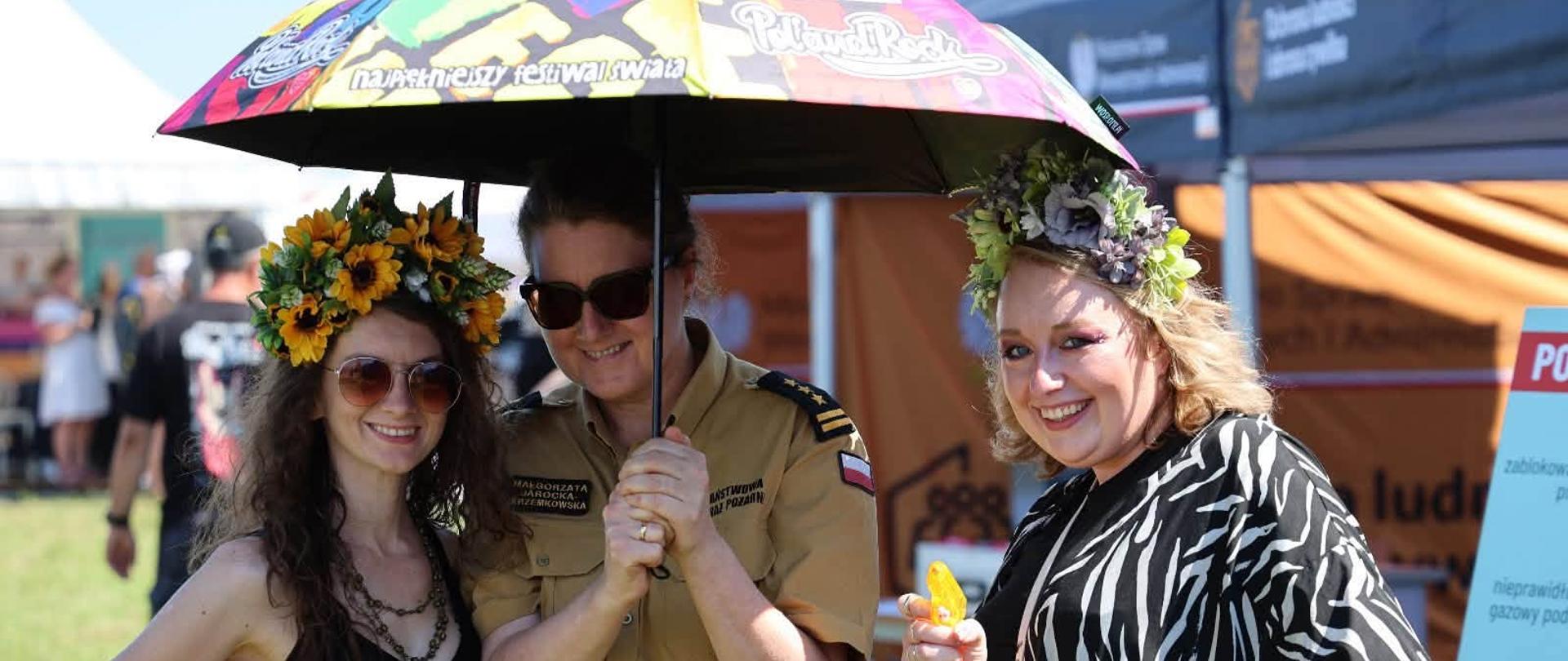 Trzy uśmiechnięte kobiety pozują do zdjęcia pod kolorowym parasolem Pol’and’Rock Festival. Kobieta pośrodku nosi beżowy mundur Państwowej Straży Pożarnej, a towarzyszące jej uczestniczki mają na głowach kolorowe wianki z kwiatów. W tle widoczne są namioty edukacyjne służb ratowniczych.