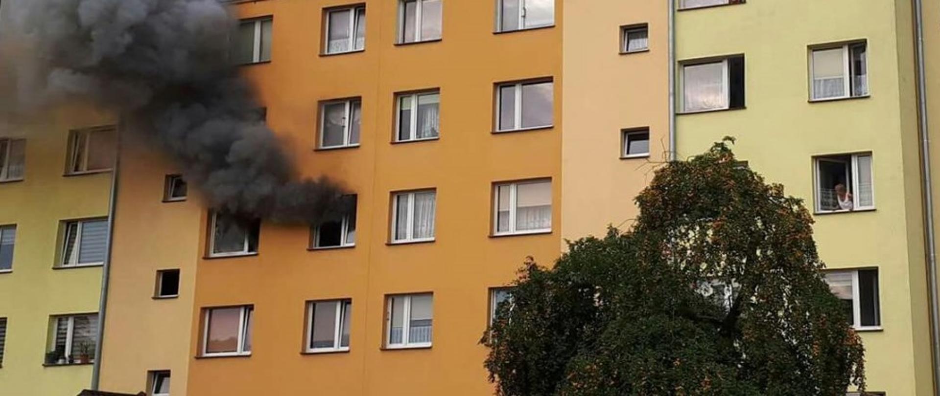 Zdjęcie przedstawia zewnętrzną ścianę budynku wielorodzinnego. Z jednego z okien budynku widać wydobywający się dym