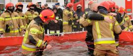 Czterech strażaków emerytów, stojących w basenie z zimną wodą, w tle oklaskujący ich przyjaciele i wóz strażacki.