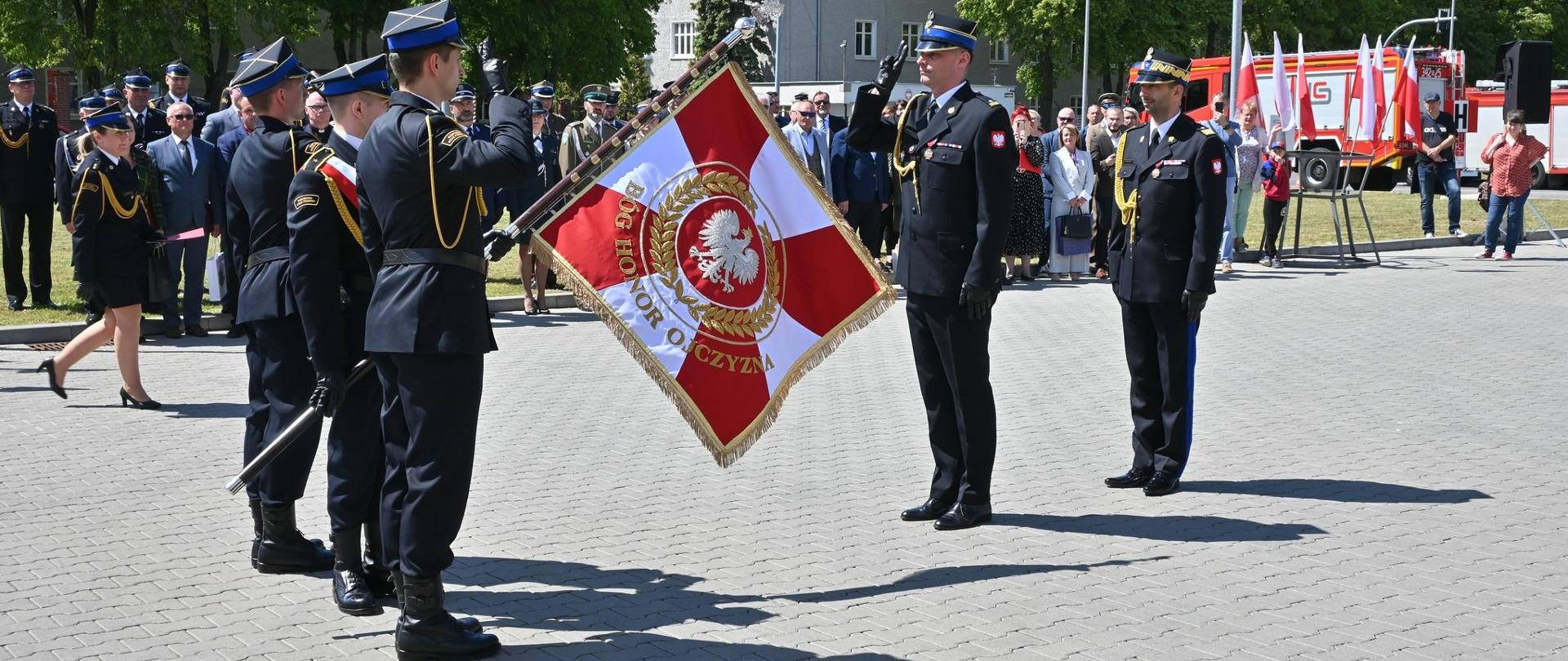 Na placu przed Komendą Miejską stoją zgromadzeni goście oraz strażacy. Na środku placu Warmińsko Mazurski Komendant Wojewódzki stoi wraz z Komendantem Miejskim na przeciw pocztu sztandarowego. W tle widoczne zabudowania i pojazdy pożarnicze.