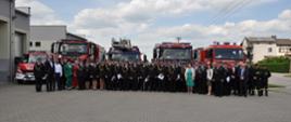 Powiatowe Obchody Dnia Strażaka 2023 r. - strażacy i goście stoją na zbiórce przed strażnicą na placu. za nimi stoją samochody strażackie