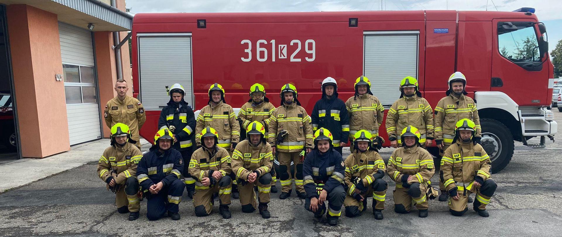 Druhowie po zakończonym egzaminie praktycznym w ubraniach specjalnych koloru piaskowego i czarnego ustawieni są w dwóch szeregach na tle samochodu pożarniczego
