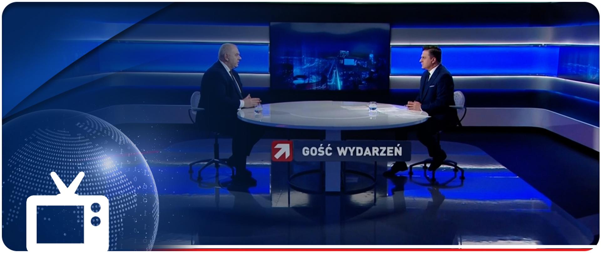 Screen programu w TV. Szerokie ujęcie studia. Przy stole siedzą: prowadzący Bogdan Rymanowski i wicepremier Jacek Sasin. Pod stołem napis Gość Wydarzeń. Na obrazek nałożony piktogram odbiornika telewizyjnego.